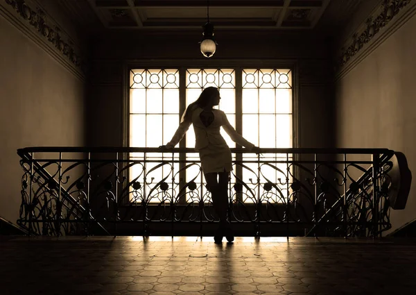 Woman in short skirt near iron stairs. Portrait full length of beautiful young adult slim aucasian woman standing against old window.