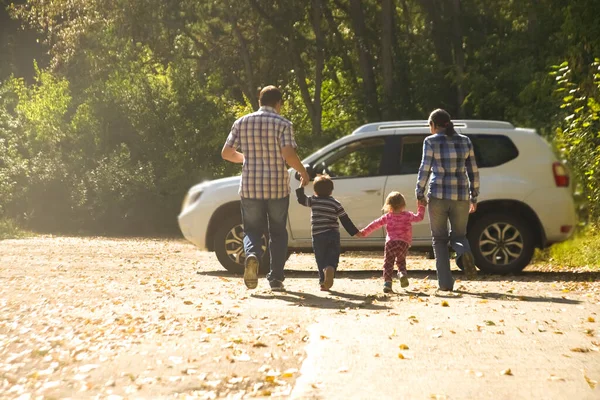 mother, father and kids - daughter, son holding hands  against white SUV big car. Outdoor photo in autumn park. Casual dress. Sunset light rays