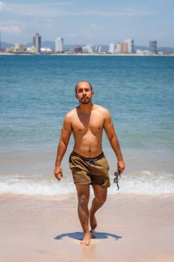 Genç Latin adam sahilde elinde güneş gözlüğü olan kameraya bakıyor.