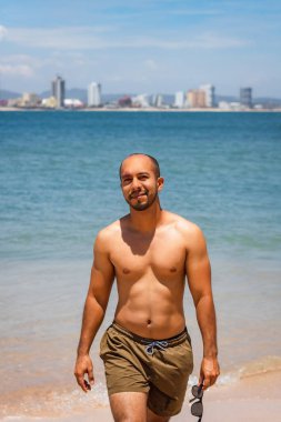 Plajda gülümseyen güneş gözlüklü genç Latin bir adam kameraya bakıyor.