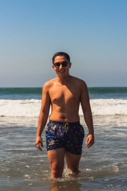 Genç Latin, gülümseyen bir adam. Denizde güneş gözlüğü takmış, yazın kameraya bakıyor.