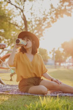 Arka planda altın saat bisiklet süren parkta su içen genç bir kadın var.