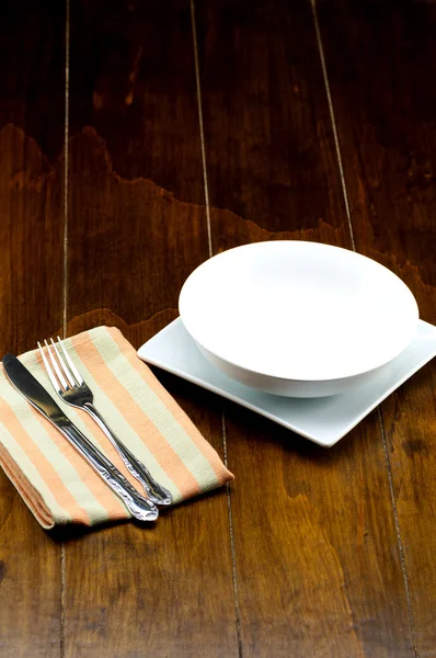 Mangkuk kosong pada piring persegi dengan garpu dan pisau di napery, di latar belakang kayu . — Stok Foto