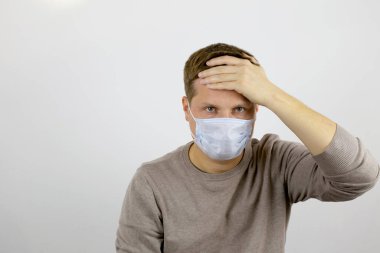 Tıbbi maskeli, başı ağrıyan bir adam kameraya bakar ve alnını eliyle tutar..