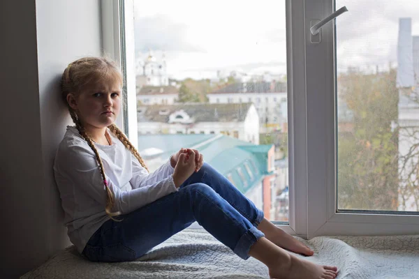 Una chica disgustada y triste se sienta en el alféizar de la ventana y mira furiosamente a la cámara. —  Fotos de Stock