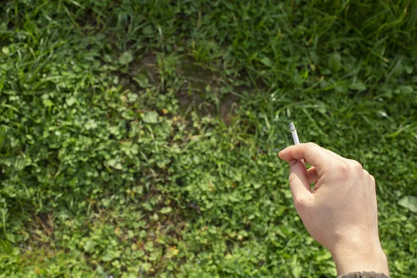 Una mano de hombre sostiene un cigarrillo contra un fondo de hierba verde brillante. —  Fotos de Stock