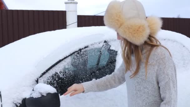 En flicka i pälsmössa och tröja rengör bilen från snö. — Stockvideo