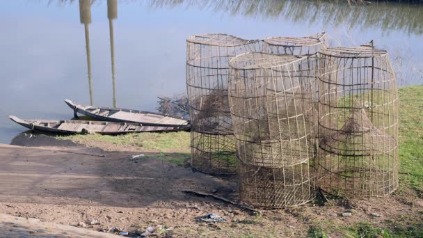 Gros plan des pièges à poissons en bambou sur la rive ; Petits pirogues amarrées au bord de la rivière en arrière-plan — Video