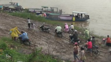 Yağmurda çamurlu bir rıhtımda motosiklet ve motosikletleriyle yürüyen feribot yolcuları.