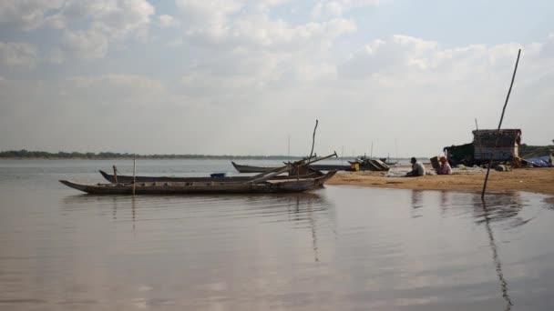 川岸に座り、手で漁網を修復する静止したダグアウトカヌーや漁師への川沿いの眺め — ストック動画