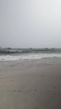 Sisli bir günde, dalgalar kumlu bir kumsala yuvarlanır ve birkaç tekne uzaktaki denize dağılır..  