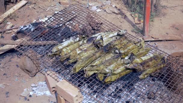 Vendedor revolviendo un fuego bajo barbacoa improvisada con pasteles de arroz de plátano cocinando en la parrilla — Vídeos de Stock