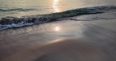 Gün batımında kumlu bir plaja doğru yavaşça süzülen köpüklü dalgalar. (Ağır çekimde ) 