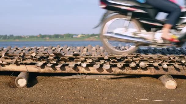 Motocicletas que viajan a través de puente de bambú / suelo colocado sobre arena inundada a lo largo de la orilla del río — Vídeos de Stock