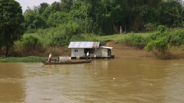 Houseboat at the river 's edge and tropical forest around; Small fishing boat passing by — стоковое видео