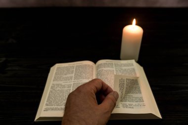 Bir mumun yanında duran alevin sıcak ışığıyla aydınlatılan koyu ahşap arka planda duran bir İncil. Erkek el, kitap kartını çevirir..