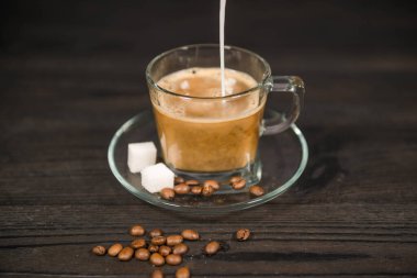 İçi demlenmiş kahveyle dolu cam bir fincan üzerinde duran şeffaf bir bardak. Kahveye yukarıdan beyaz süt dökülüyor. Bardağın yanında, dağınık kahve ve şeker küpleri var..