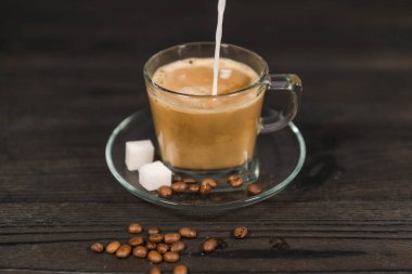 İçi demlenmiş kahveyle dolu cam bir fincan üzerinde duran şeffaf bir cam bardak. Kahveye yukarıdan beyaz süt dökülüyor. Bardağın yanında kahve çekirdekleri ve şeker küpleri var..