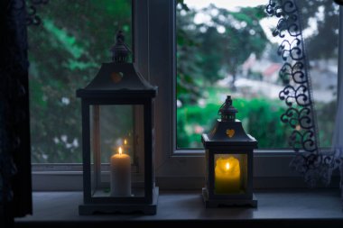 Dairenin pencere eşiğinde. En üstte bir perde görebilirsiniz, daire hem içerisi hem de dışarısı loş. Pencere pervazında iki fener var. Fenerlerin içindeki mumlar kırılmış..