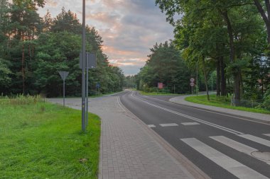 Kavisli asfalt bir yol iki enine yol geçiyor. Ağaçların arasındalar. Ağaçların üzerinde akşam kızıl gökyüzünü görebilirsin..