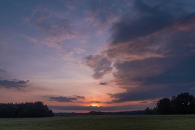 Otla kaplanmış düz bir arazi. Uzakta bir sıra ağaç var. Gökyüzü kısmen koyu bulutlarla kaplıdır. Batan güneşin ışığıyla kırmızıya boyanmıştır..