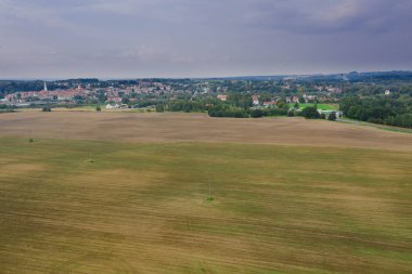 Geniş bir düzlük, yollarla geçen tarlalar ve çayırlar. Yeşil ağaç kümeleri görünür. Uzaktan yakındaki kasabanın binalarını görebilirsin. Gökyüzü hafifçe bulutlu. Fotoğraf İHA kullanılarak çekildi..