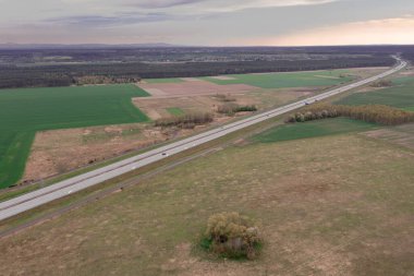 Tarlalarla, çayırlarla ve ağaçlarla kaplı bir ovanın içinden bir yol geçiyor. Güneşli bir gün. Fotoğraf İHA kullanılarak yüksek irtifada çekilmiş..