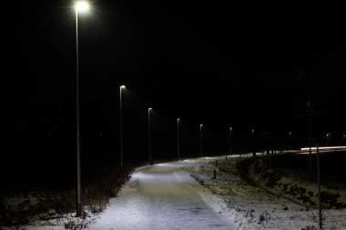 Ana yolun yanında bir bisiklet yolu. Kış mevsimindeyiz ve yüzey beyaz kar tabakasıyla kaplı. Gece oldu ve bir sıra LED lamba karanlığı aydınlatıyor. Işıkları düşen karı ortaya çıkarıyor..