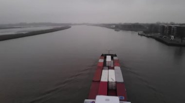 Bir yolcu feribotu sisli bir günde nehrin ters tarafından diğer tarafa doğru yol alırken, konteyner mavnası Rotterdam yakınlarındaki bir Hollanda nehrinde yol alıyor. Drone dolly yavaş çekim.