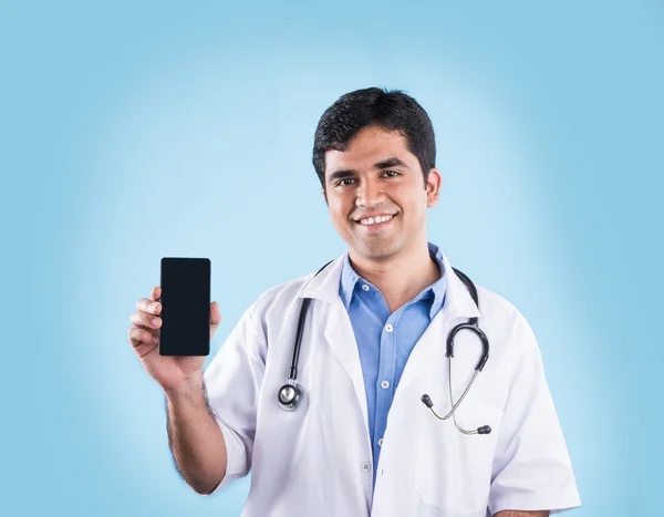 Portrait d'un médecin indien confiant montrant un téléphone intelligent sur fond bleu. médecin indien avec téléphone intelligent, asiatique médecin pointant téléphone intelligent — Photo