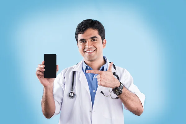 Portrait of confident indian male doctor showing smart phone over blue background. indian doctor with smart phone, asian doctor pointing smart phone — Stock Photo, Image