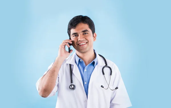 青い背景の上にスマートフォンを示す自信のあるインドの男性医師の肖像画。スマートフォンを持つインドの医師、スマートフォンで話すアジアの医師 — ストック写真