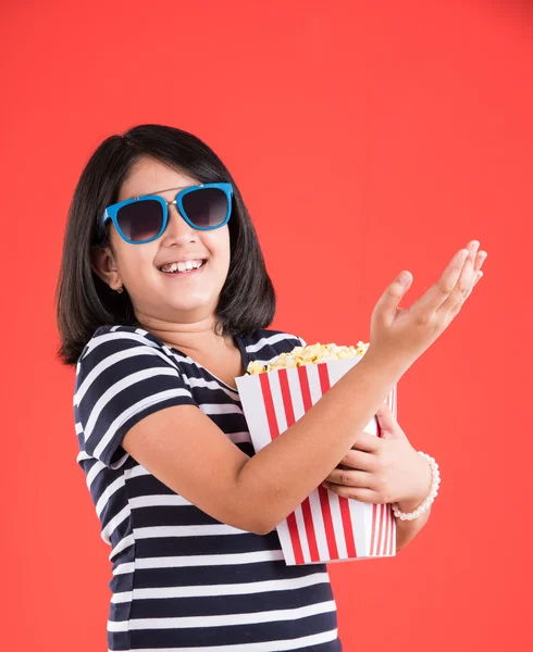Happy girl eating popcorn and wearing glasses, indian girl eating popcorn, asian girl and popcorn, small girl eating popcorn on red background — Stock Photo, Image