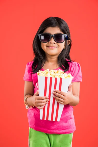 Szczęśliwy dziewczyna jedzenia popcorn i noszenie okularów, Indian Girl jedzenia popcorn, Asian Girl i popcorn, mała dziewczynka jedzenia popcorn na czerwonym tle — Zdjęcie stockowe