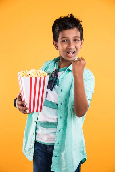 Iloinen intialainen poika, jolla on iso laatikko popcornia ja joka katsoo keltaisella taustalla eristettyä kameraa, Suloisen intialaisen pojan muotokuva, joka syö popcornia, aasialaista lasta ja popcornia. — kuvapankkivalokuva