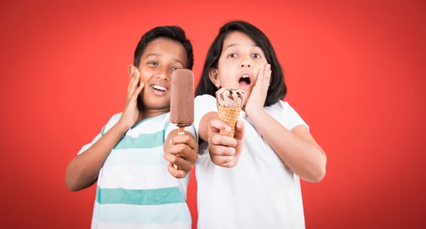 Duas crianças indianas felizes e sorvete, duas crianças asiáticas desfrutando de sorvete ou cone ou chocolate doce, menina e menino comendo sorvete, isolado no fundo vermelho , — Fotografia de Stock