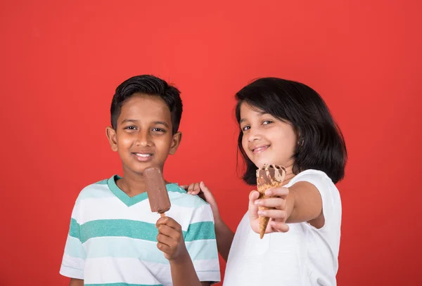 Two happy indian kids and ice cream, two asian kids enjoying ice cream or cone or chocolate candy, girl and boy eating ice cream, isolated on red background, — Stock Photo, Image