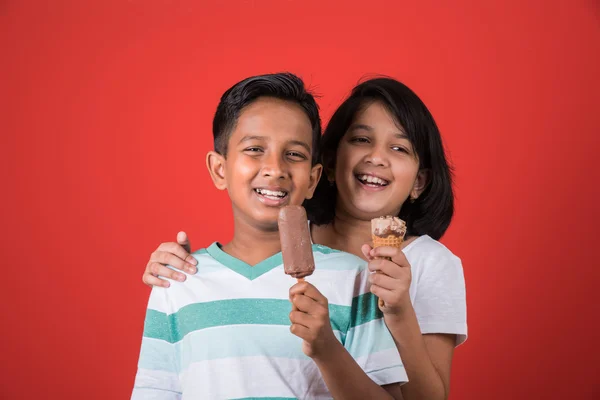 Két boldog indiai gyerekek és fagylalt, két ázsiai gyerekek élvezik fagylaltot, vagy kúp vagy csokoládé cukorka, lány és fiú étkezési fagylalt, elszigetelt vörös háttér, — Stock Fotó