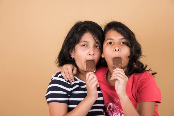 two indian girls eating ice cream or ice candy, two asian girl and ice cream or ice candy, isolated on brown background, ten year old indian girls enjoying ice cream or ice candy or choko bar