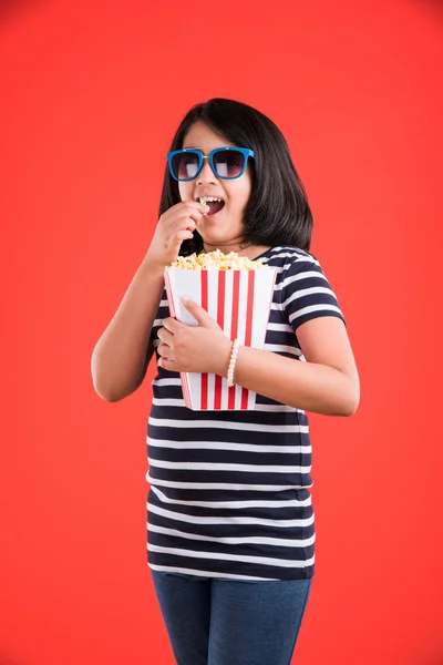 happy girl eating popcorn and wearing glasses, indian girl eating popcorn, asian girl and popcorn, small girl eating popcorn on red background