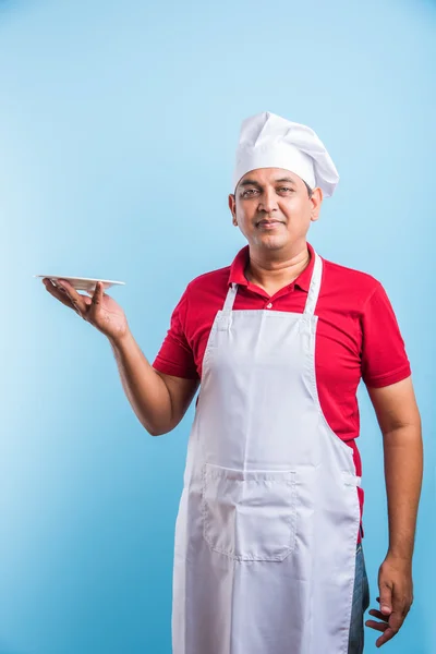 Portrait of an Indian Male cook in uniform holding an empty dish, presenting something. standing isolated over blue background, asian chef holding red plate — Stock Photo, Image
