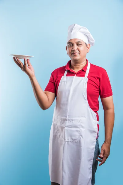 Портрет індійського чоловічого кухаря в уніформі тримає порожню страву, представляючи щось. Постійна ізольовані над синім фоном, Азіатський шеф-кухар тримає червону пластину — стокове фото