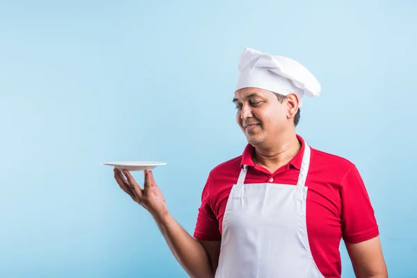 Retrato de um cozinheiro indiano macho de uniforme segurando um prato vazio, apresentando algo. pé isolado sobre fundo azul, asiático chef segurando placa vermelha — Fotografia de Stock