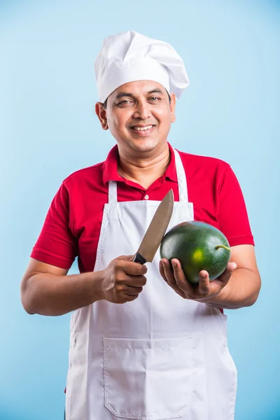 Індійський чоловічий шеф-кухар тримає ніж в одній руці і фрукти дині з іншого боку, Азіатський чоловічий шеф-кухар позує з великим ножем і кавун, ізольовані над синім фоном — стокове фото