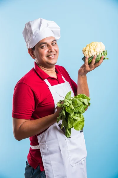 indian male chef holding fresh Cauliflower and spinach leaves, standing isolated over blue background, asian male chef holding fresh vegetables