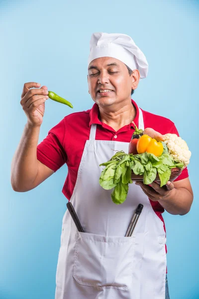 indian male chef holding fresh vegetables in hand, asian male cook chef standing with vegetables, isolated over blue background