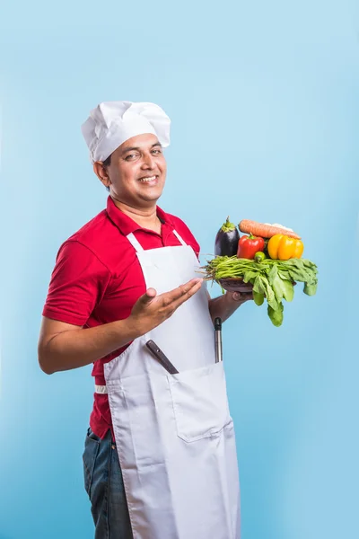 indian male chef holding fresh vegetables in hand, asian male cook chef standing with vegetables, isolated over blue background