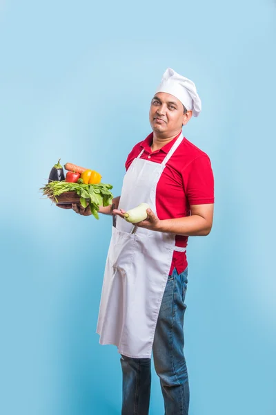 indian male chef holding fresh vegetables in hand, asian male cook chef standing with vegetables, isolated over blue background