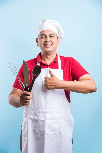 Multitasking is a chefs game. Closeup portrait of handsome indian cook holding a wooden culinary spoon and soup ladle and knife while standing over blue background with copy space, asian male chef — Stock Photo, Image