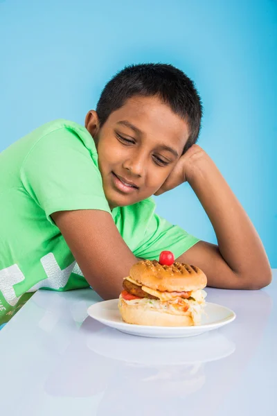 Schattige Indiase jongen en Hamburger in kleine witte plaat, kleine Aziatische jongen en Hamburger, over kleurrijke achtergrond — Stockfoto